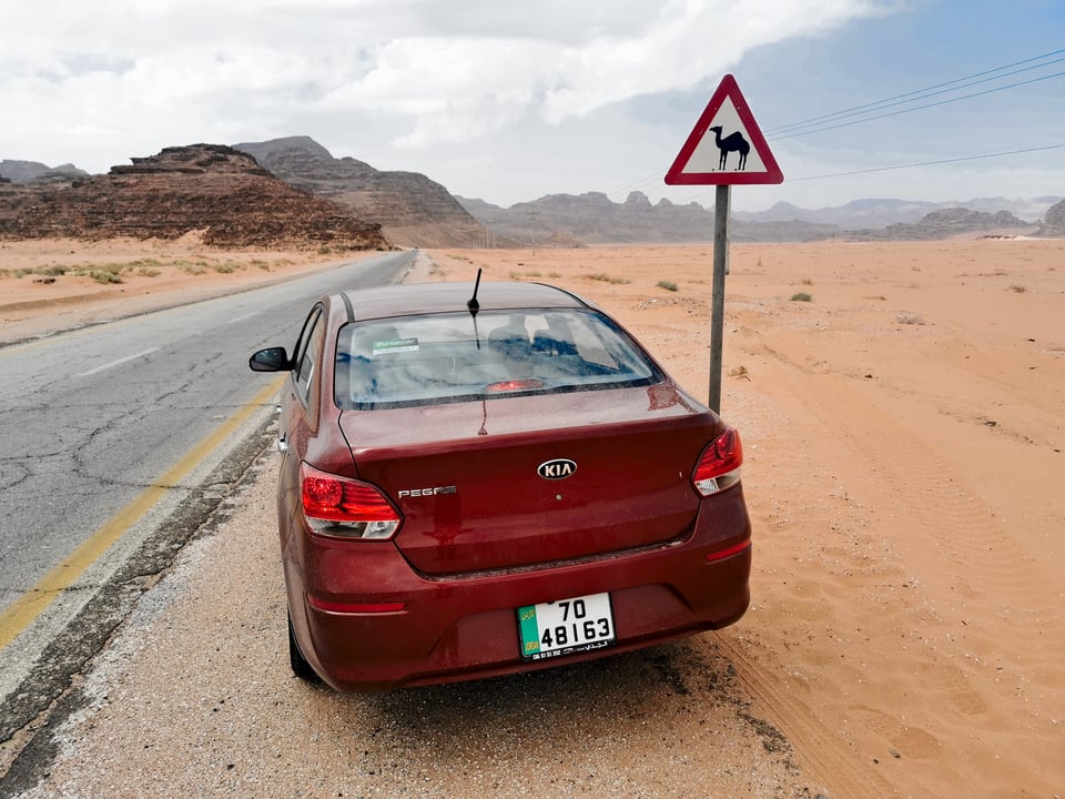 Een rode huursedan langs een zanderige woestijnweg in Jordanië, naast een waarschuwingsbord voor overstekende kamelen, met rotsbergen op de achtergrond.